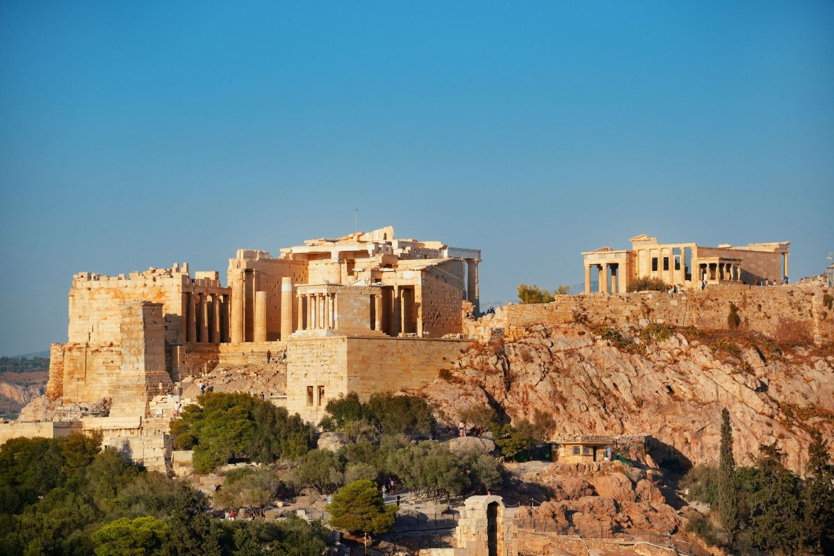 The image shows the Acropolis of Athens, featuring the Parthenon and other ancient Greek structures on a rocky hill. The scene includes a clear blue sky and surrounding greenery.